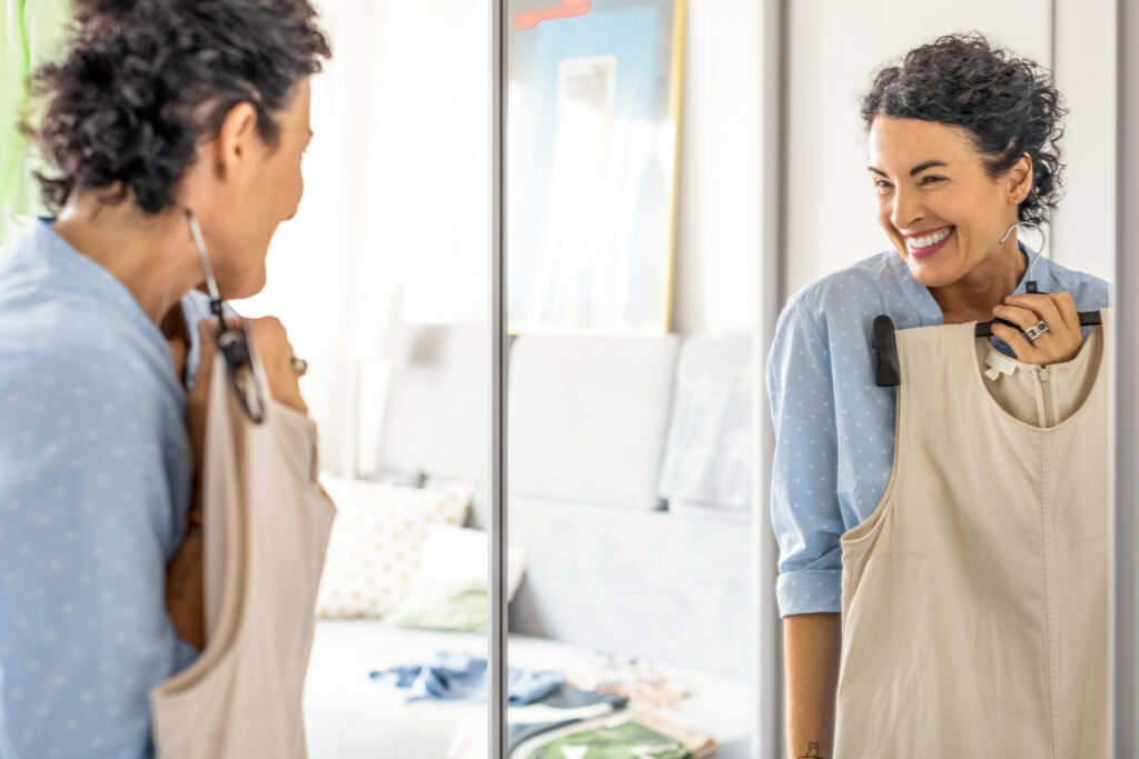 Frau 50+blickt in den Spiegel mit Kleidung in der Hand